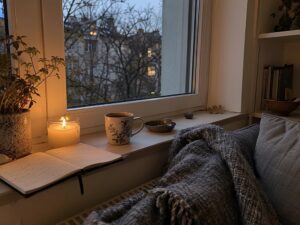 Fensterbank mit Teetasse, Kerze, Notizbuch und Wolldecke in der herbstlichen Abenddämmerung